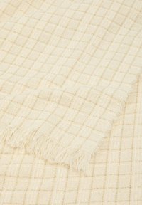 Coperta in crema con un motivo a griglia testurizzato e fili dorati. I bordi rifiniti sono frangiati, accentuando il dettaglio tattile. Morbida e leggera.