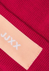 Tessuto lavorato a maglia rosso con un aspetto testurizzato. Etichetta color pesca chiaro con testo bianco "JJXX." Design semplice con colori a contrasto.