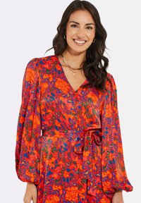 Bunt gemustertes Kleid mit V-Ausschnitt, langen Puffärmeln und einer gebundenen Taille. Es hat ein lebhaftes Blumen- und Tiermuster in Rot-, Blau- und Grüntönen.