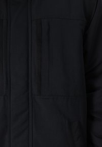 Black jacket with a matte finish, featuring two chest pockets, horizontal seam details, and concealed zippers for a streamlined aesthetic.