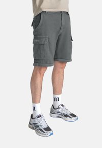 Shorts cargo gris en tissu solide, avec deux poches latérales, des ourlets roulés et une coupe décontractée. Portés avec des chaussettes blanches et des baskets.
