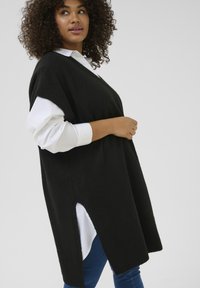 Czarny oversize sweter kamizelkowy z miękkiej przędzy, z dekoltem w serek, bocznymi rozcięciami i ribbendingiem. Nałożony na białą koszulę, noszony z niebieskimi dżinsami.