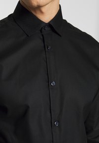 Camicia nera a maniche lunghe realizzata in tessuto liscio, con colletto a punta e bottoni frontali di tonalità scura, ben stirata e senza fantasie.