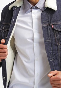 Giacca di jeans scura con una fodera in pile bianco, indossata sopra una camicia grigio chiaro con bottoni. Presenta bottoni in metallo e cuciture a contrasto.