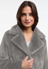 Grijze nepbontjas met een brede kraag, zachte textuur en verlengde silhouet. Het model houdt de jas gesloten bij de borst.