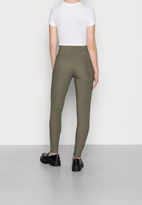 Osoba ubrana w oliwkowe legginsy o wysokim stanie, białą, krótką bluzkę z krótkim rękawem oraz czarne, masywne mokasyny, stojąca przy białej ścianie.