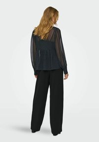 Femme aux longs cheveux blonds portant un chemisier noir transparent à pois et un pantalon noir à jambes larges, debout de dos sur un fond uni.