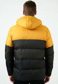 Puhasta jakna z rumenim hoodiejem in črnim telesom, ki ima prešito zasnovo in toplo, obloženo teksturo. Primerna za hladno vreme.