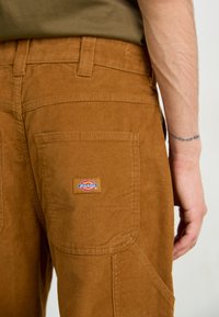 Pantaloni din catifea marron cu un buzunar pe partea dreaptă, având un logo Dickies roșu și albastru.Țesătură texturată și detalii de cusătură vizibile.