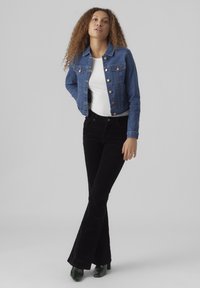 Denim jakna u plavoj boji, s bakrenim gumbima i dva džepa na prsima, u kombinaciji s crnim korduroy širokim hlačama i zelenim potpeticama. Ispod se nalazi bijela košulja.