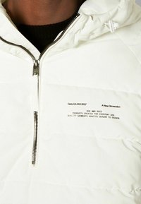 Vit puffjacka med dragkedja framtill, med horisontella delade sektioner och svart tryckt textdetalj på vänster bröst.