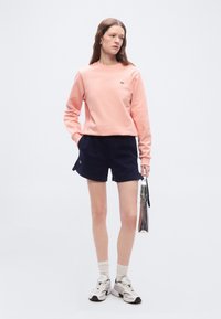 Νεαρή γυναίκα φοράει ροζ φούτερ, ναυτικό σορτς, λευκά αθλητικά παπούτσια και κρατάει μια μικρή ασημί τσάντα χειρός μπροστά σε λευκό φόντο.