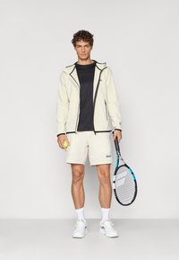 Lys cremet zip-up jakke, sort t-shirt, off-white shorts med logo, holder en tennisbold og ketsjer, hvide sneakers, krøllet hår.