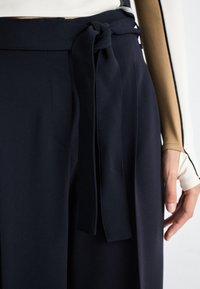 Pantaloni blu navy dalla silhouette ampia con cintura in tessuto legata. Presentano una texture liscia e tasche laterali. Il materiale è leggero e si drappeggia morbidamente.
