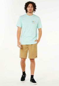 Helles blaues Baumwoll-T-Shirt mit Frontgrafik, beigefarbene Cordshorts und schwarze Sneaker. Model steht mit den Händen in den Taschen vor einem neutralen Hintergrund.