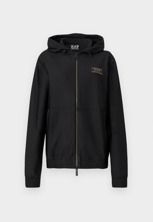 Veste noire à fermeture éclair avec tissu texturé, capuche attachée, poignets élastiques et taille élastique. Détail logo doré sur la poitrine.