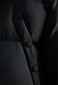 Detail čiernej puffovej bundy s hladkým povrchom, zapínaním na gombíky s vyrytým logom a prešívaným dizajnom, ktorý zdôrazňuje vypchávkové sekcie.