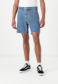 Shorts din denim de culoare albastră deschisă, croială normală, margine desfăcută, cinci buzunare, închidere cu nasture, purtate cu adidași negri și albi și șosete albe până la glezne.