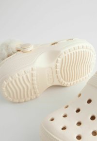Des chaussures slip-on beiges claires avec une semelle texturée, de grands orifices de drainage et un accent en fourrure douce près de la fermeture à bouton.