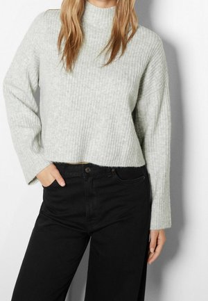 Femme portant un pull en maille côtelée gris clair avec un col haut et un pantalon noir taille haute, main dans la poche, sur un fond uni.