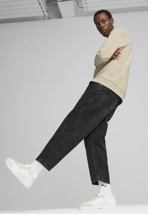 Beiger Sweatshirt, schwarze lockere Jeans und weiße Sneakers mit beigen Akzenten. Die Socken sind weiß mit einem Logo. Stehende Pose mit einem Bein angehoben.