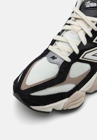 Gros plan d'une basket New Balance 9060 noire, beige et blanche avec des détails en mesh et en daim et des lacets couleur crème.