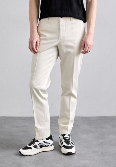 Pantaloni crem, croiți, dintr-un material ușor, având un finisaj neted și un design cu picioare drepte. Asortați cu adidași negri și albi.