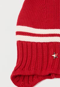 Czerwona dzianinowa czapka beanie o prążkowanej strukturze, z dwoma białymi paskami oraz małą przypinką w kształcie gwiazdki w kolorze czarnym, czerwonym i białym.