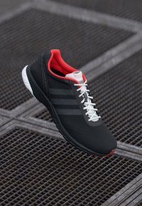 Black athletic shoe with red accents, grey stripes, and white laces. Features a textured upper and a sporty, streamlined design.