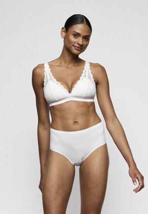 Vrouw met donker haar in een witte kanten beha en witte hoge tailleonderbroek staat voor een effen lichte achtergrond, kijkt zacht lachend.