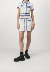 Witte cropped gebreide top met blauwe randen, korte mouwen en knopen; bijpassende witte mini rok met blauwe accenten en zakken. Zwarte platform schoenen.