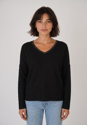 Mujer con el cabello corto y oscuro, vestida con un suéter negro de manga larga con cuello en V y vaqueros azul claro, de pie frente a un fondo liso y claro.