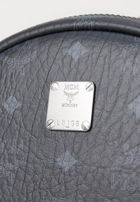 Hall sinine tekstureeritud nahk ja hõbedane metallplaat, millel on brändi nimi "MCM" ja reljeefsed detailid; sisaldab riistvara aktsente.