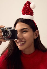 Woman wearing a red sequined Christmas hat, holding a vintage black camera with silver accents, against a neutral background. Red fuzzy sweater visible.