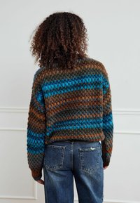 Kootud kampsun, millel on triibud pruunis, sinises ja rohelises toonis, kombineeritud kõrge vöökohaga siniste teksadega. Tekstureeritud lõng ja lõtv lõige.