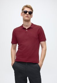 Burgundy polo shirt with short sleeves, collar, and two buttons, worn with dark gray trousers. Model wearing black sunglasses.