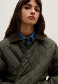 Junge Frau mit langen braunen Haaren, die eine dunkelgrüne gesteppte Jacke und ein blaues Hemd mit Kragen trägt, vor einem neutralen Hintergrund.