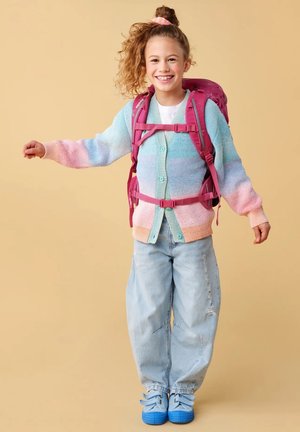 Smiling girl with curly hair in a pastel cardigan, ripped jeans, blue shoes, and pink backpack straps, standing against beige background.