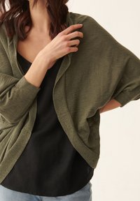 Olijfgroene open voorkant cardigan gemaakt van een textuurstof, gelaagd over een zwarte top. De cardigan heeft brede mouwen en een ontspannen pasvorm.