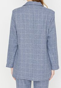 Blazer gris sur mesure avec motif à carreaux blancs, fabriqué en tissu texturé, doté d'un col cranté et de manches longues. Détail discret de fente au dos.