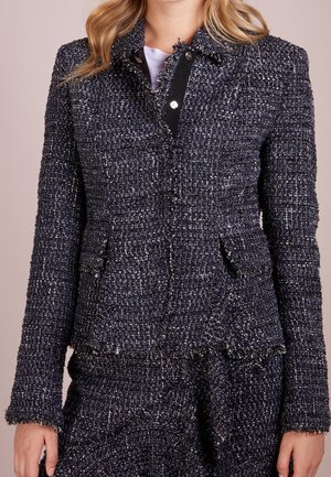 Vrouw draagt een donker tweed-textuur blazer met bijpassende rok, met klepzakken en rafelige randen over een witte top.