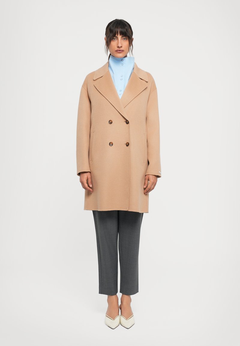 Donna in piedi che indossa un cappotto cammello doppiopetto, una camicia colletto alto azzurro chiaro, pantaloni grigi e scarpe bianche a punta su sfondo bianco.