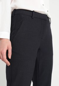 Tailored black trousers with a smooth texture, featuring side pockets and a slim fit, shown alongside a white blouse.