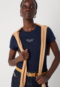 Marineblå bomull T-skjorte med brodert logo, kombinert med en lys beige genser knyttet rundt skuldrene, og et gult belte på mørke jeans.