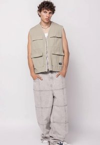 Smėlio spalvos rankovių liemenė su priekinėmis kišenėmis ir užtrauktuku, dėvima ant baltos marškinėlių su petnešėlėmis, derinama su pilkomis plačiomis kelnėmis su siuvinėjimo raštais.