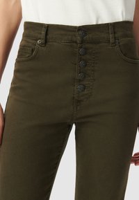 Nabijend van een persoon die hoogwaardig, donkergroen broek draagt met vijf zichtbare zwarte knopen en voorzakken, gecombineerd met een wit topje.