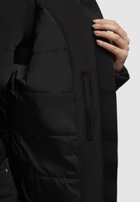 Black insulated jacket with smooth exterior, padded interior, and a side hand pocket. Stitched detailing enhances structure and design.