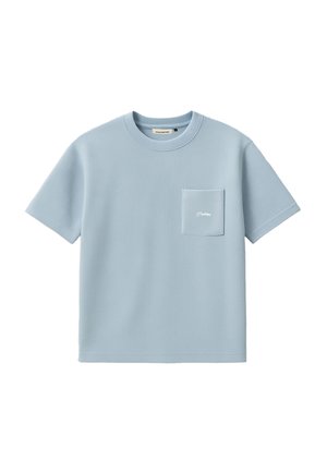 Giovane uomo che indossa una maglietta azzurro chiaro oversize, una sciarpa nera paisley al collo, pantaloni neri e un berretto beige con scritta rossa e blu.