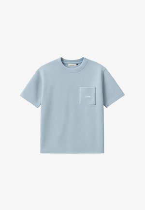 Ung mand iført en lyseblå oversized t-shirt, sort paisley halsrør, sorte bukser og beige kasket med rød og blå tekst.