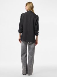 Eine Frau mit langen blonden Haaren trägt ein schwarzes Nadelstreifen-Blazer mit hochgekrempelten Ärmeln und weiten, hellgrauen Jeans, sie steht vor einem weißen Hintergrund.